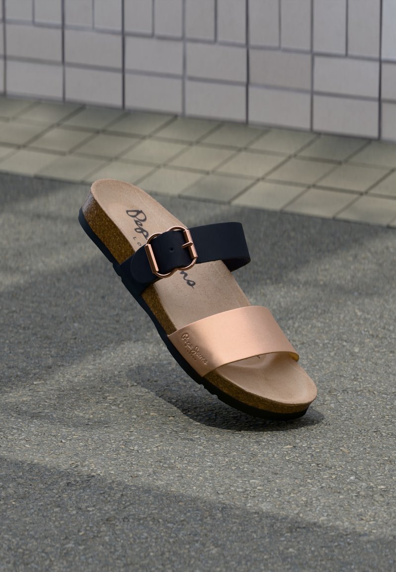 Σανδάλι με φελλώδη σόλα, μαύρο ρυθμιζόμενο λουράκι με αγκράφα και μεταλλικό λουράκι μπροστά σε ροζ χρυσό σε γκρι πλακόστρωτο φόντο.