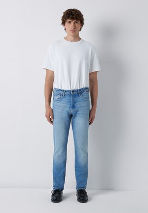 Young man with curly hair wearing a plain white t-shirt, light blue jeans, and black shoes standing against a white wall.