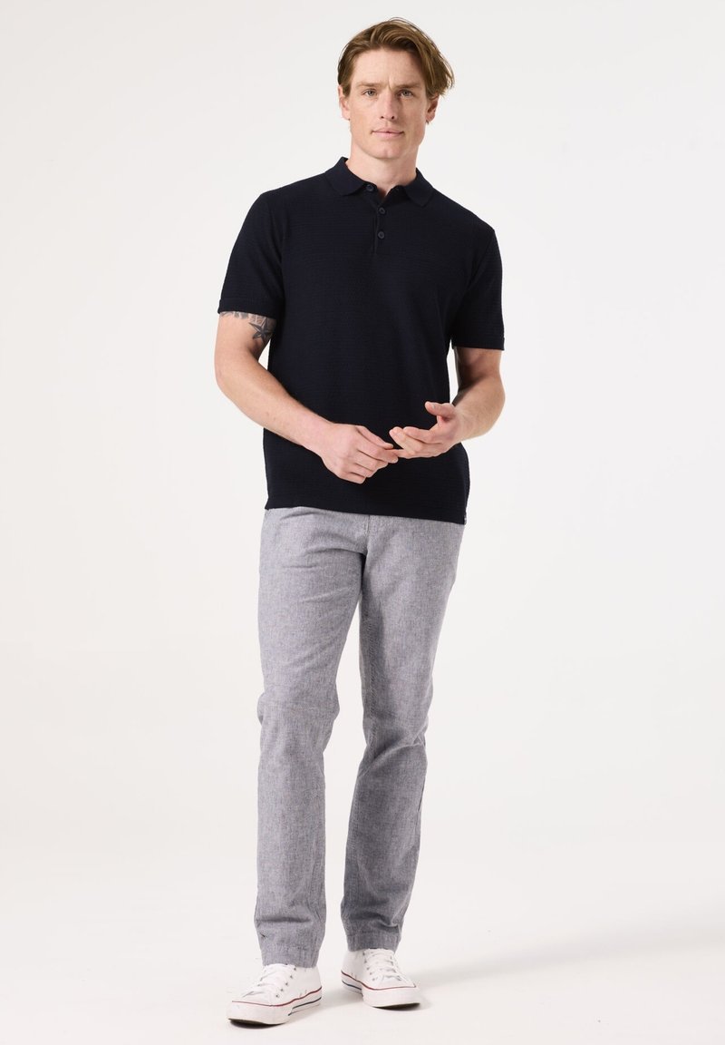 Marineblauw polo-shirt met korte mouwen en een gestructureerd brei-design, gecombineerd met lichtgrijze broek en witte sneakers, staand tegen een effen achtergrond.