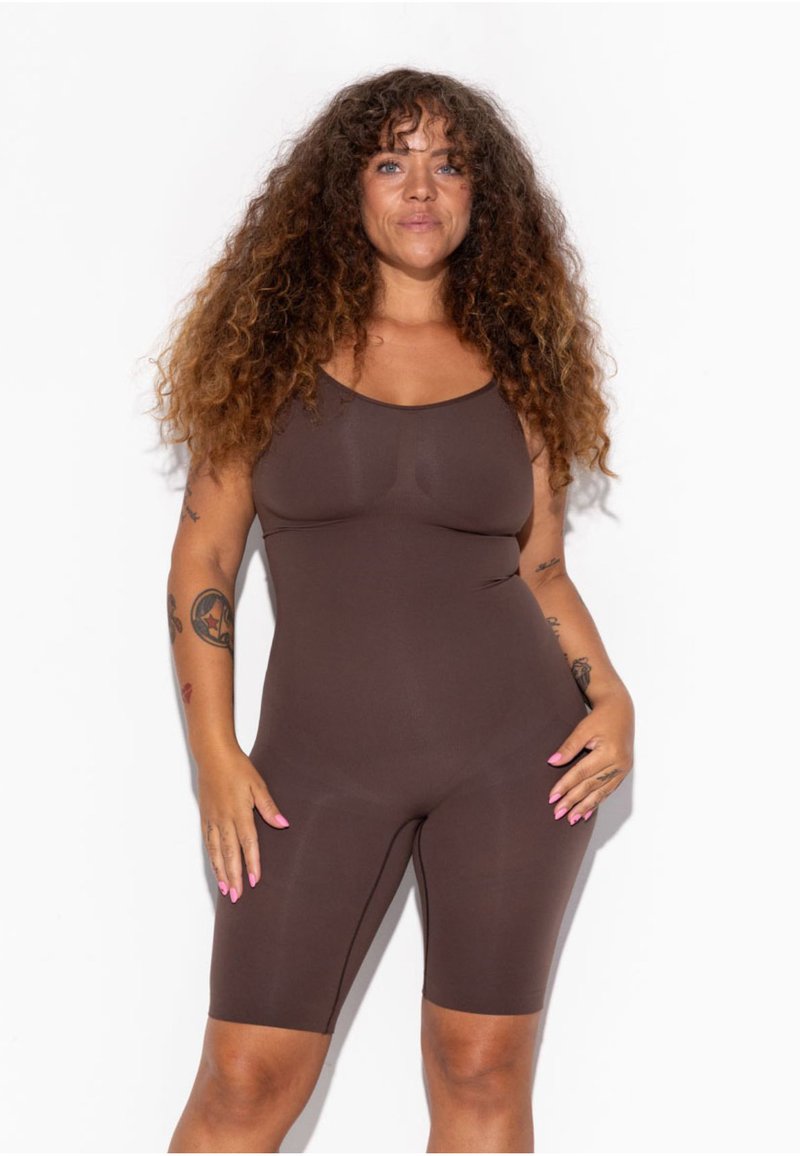 Femme aux longs cheveux bouclés portant une combinaison marron moulante sans manches, debout devant un fond blanc avec des tatouages visibles sur les bras.