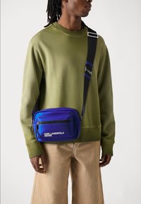 Blå gradient crossbody veske med en glidelåslomme på fronten, innebygd logo, justerbar stropp og rund rektangulær form. Teksturert materiale.