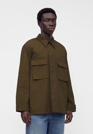 Homme portant une chemise boutonnée vert olive oversize avec de grandes poches à rabat et un jean bleu sur un fond uni.
