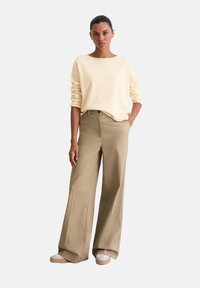 Hellgelbes Sweatshirt mit aufgekrempelten Ärmeln, kombiniert mit weiten beigen Hosen. Die Schuhe haben eine helle Sohle und einen braunen Akzent.