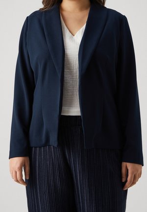Femme portant un blazer bleu marine sur un haut blanc texturé à col en V et un pantalon plissé bleu marine, debout devant un fond clair uni.