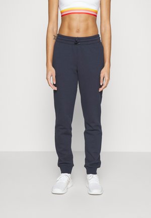 Person iført mørkeblå joggers, hvide sneakers og en hvid crop top med røde, orange og gule striber på en ensartet lys baggrund.