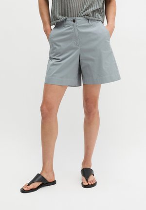 Persoon draagt lichtgrijze knielange shorts, een grijs mouwloos gebreid topje en zwarte instap sandaaltjes, staand met handen in de zakken.