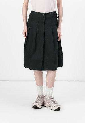 Jupe plissée noire avec une fermeture à boutons, texture du tissu visible ; modèle associé à des chaussettes blanches aux chevilles et des baskets de couleur claire.