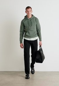 Sudadera verde con "GANT" en relieve, combinada con una camisa blanca, pantalones negros y un bolso negro texturizado. Modelo llevando zapatos negros, caminando.