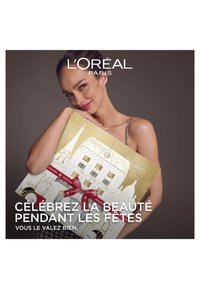 Boîte-cadeau ornée d'un design doré avec des illustrations festives et des rubans rouges, tenue par une personne portant une robe à sequins et des boucles d'oreilles.