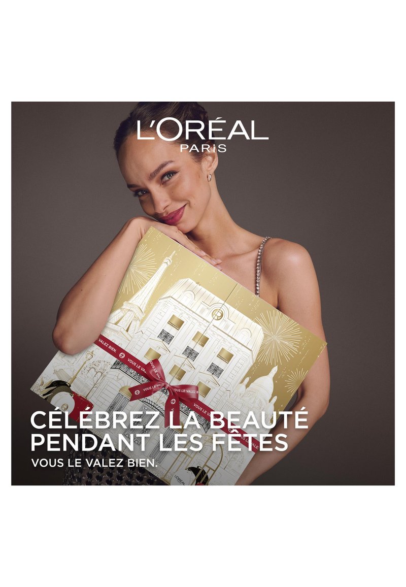 Boîte-cadeau ornée d'un design doré avec des illustrations festives et des rubans rouges, tenue par une personne portant une robe à sequins et des boucles d'oreilles.