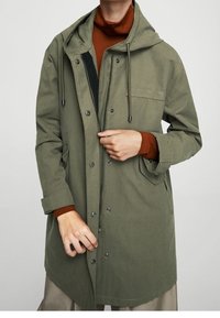 Parka olive verte avec capuche, fermetures à pression sur le devant et poches latérales. Présente une coupe décontractée et une texture de tissu lisse.
