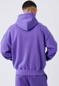 Sudadera con capucha morada, fabricada en un tejido suave, que presenta un corte relajado, bolsillo tipo canguro y puños y bajo acanalados. Se muestra vista posterior.