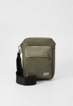 Bolso cruzado de nailon verde oliva con un bolsillo frontal, correa negra y un acento de logo metálico. Textura suave y cierre con cremallera.