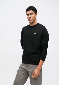 Μαύρο φούτερ με στρογγυλή λαιμόκοψη και ριμπ μανσέτες, με λευκό λογότυπο "Barbour" στο στήθος. Φορεμένο με γκρι παντελόνι.