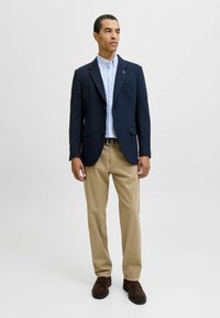 Blazer navy con dos bolsillos frontales, camisa de botones azul claro, pantalones beige y zapatos marrones. Líneas limpias y tejido texturizado.
