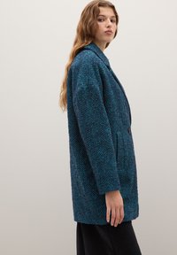 Un cappotto herringbone texturizzato in verde acqua e nero con una silhouette casual, ampi rever e chiusura a un bottone, indossato sopra pantaloni scuri.