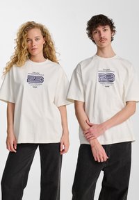 Twee personen dragen oversized beige t-shirts met een marineblauwe grafische print, gecombineerd met donkere jeans. De stof lijkt zacht en casual.