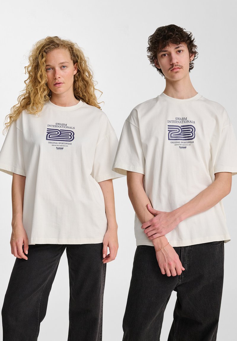 Twee personen dragen oversized beige t-shirts met een marineblauwe grafische print, gecombineerd met donkere jeans. De stof lijkt zacht en casual.