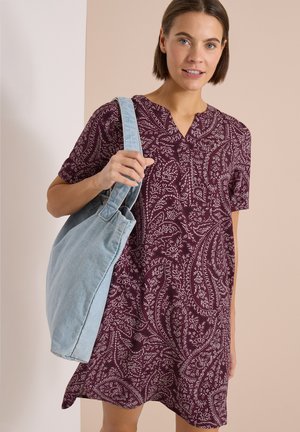 Frau trägt ein kurzärmeliges lila Paisley-Kleid und trägt eine große hellblaue Schultertasche, steht vor einem beige-weißen Hintergrund.