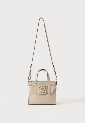 Sac à main beige à la forme structurée, avec de courtes poignées, une longue bandoulière réglable et un logo géométrique en relief, sur un fond uni.