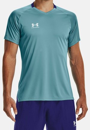 Mężczyzna w jasnoturkusowej sportowej koszulce z krótkim rękawem marki Under Armour i ciemnofioletowych szortach, stojący z rękami swobodnie opuszczonymi wzdłuż ciała.