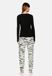 Woman with straight brown hair wearing a black long-sleeve top, white pants with black abstract stripes, and silver sneakers, facing away.