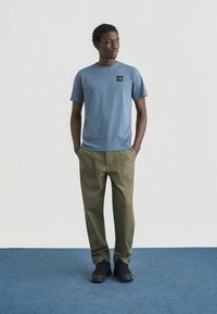 Μπλε βαμβακερό μπλουζάκι με μαύρο λογότυπο, συνδυασμένο με χακί παντελόνι. Το μοντέλο φοράει μαύρα αθλητικά παπούτσια και στέκεται σε μπλε χαλί.