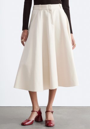 Modèle portant une jupe midi taille haute couleur crème avec des plis, associée à un haut marron foncé et des chaussures Mary Jane rouges à talons.