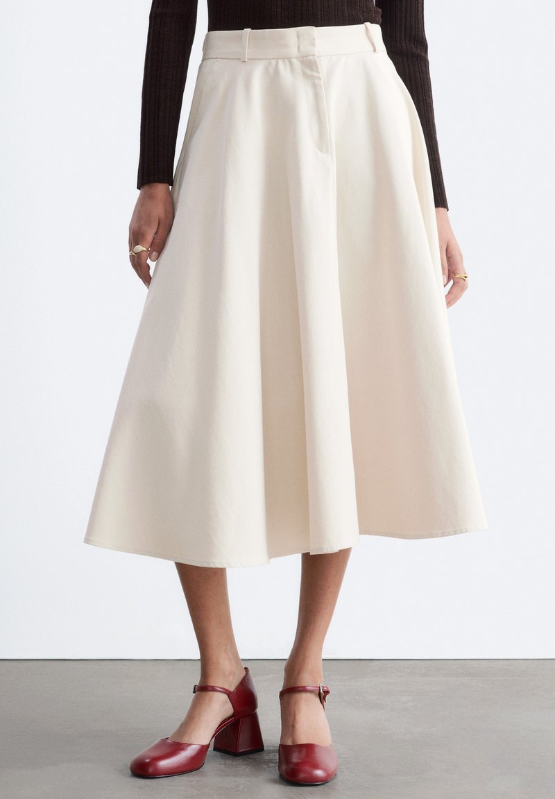 Modèle portant une jupe midi taille haute couleur crème avec des plis, associée à un haut marron foncé et des chaussures Mary Jane rouges à talons.