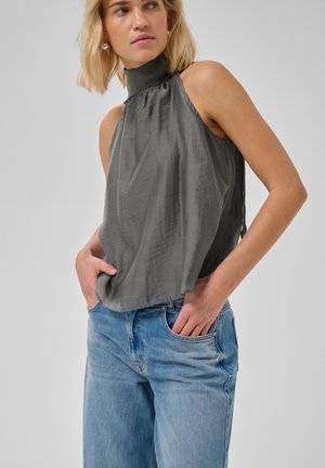 Mujer rubia que lleva una camiseta gris sin mangas de cuello alto y vaqueros azules con las manos en los bolsillos, mirando ligeramente de lado sobre un fondo liso.