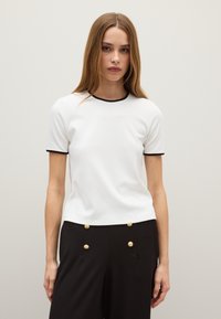 Maglietta bianca a maniche corte con bordo nero al collo e alle maniche. Tessuto morbido con un design aderente. Indossata con pantaloni neri a vita alta con bottoni dorati.