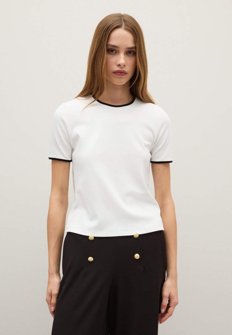 Maglietta bianca a maniche corte con bordo nero al collo e alle maniche. Tessuto morbido con un design aderente. Indossata con pantaloni neri a vita alta con bottoni dorati.