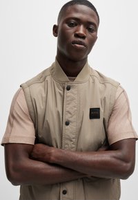 Beige ärmellose Jacke mit glatter Textur und geripptem Kragen, ausgestattet mit einem schwarzen Etikett mit "BOSS"-Logo und dunklen Knöpfen über einem hellbraunen Hemd.