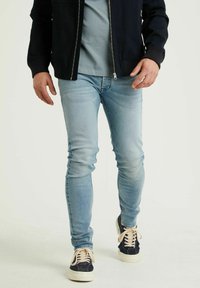 Jeans ajustados de color azul claro hechos de denim, combinados con una chaqueta oscura con cremallera y una camiseta azul, terminados con zapatillas marinas con cordones crema.