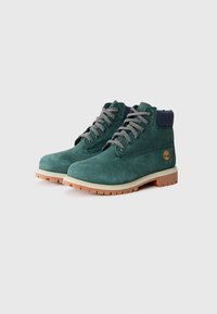 Stivaletti alla caviglia in suede verde con colletto blu scuro, lacci grigi e suola in gomma beige. Presentano il logo sul lato e una texture liscia.