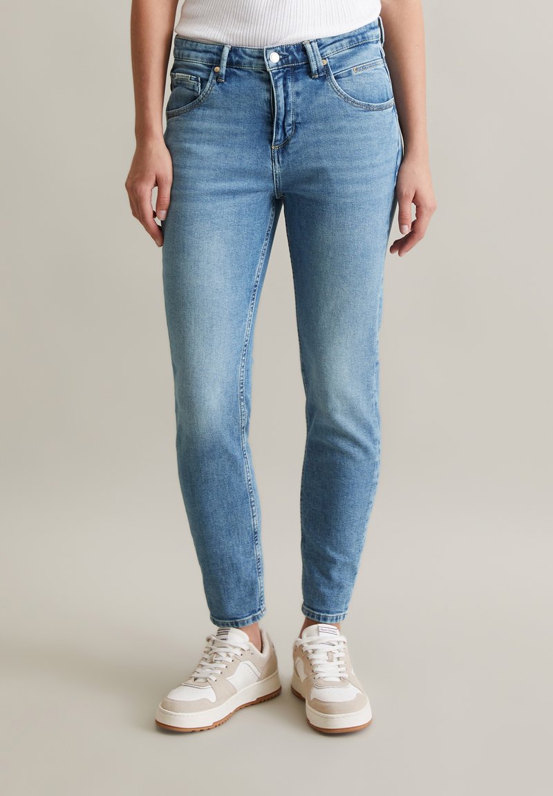 Jasnoniebieskie slim fit dżinsy o gładkiej teksturze denimu, pięć kieszeni, zapięcie na guzik i zamek błyskawiczny, w połączeniu ze kremowymi sneakersami.