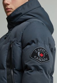 Chaqueta azul marino acolchada con cuello alto, bolsillo lateral con cremallera y un parche de logo bordado en negro con una hoja de arce roja en el hombro.