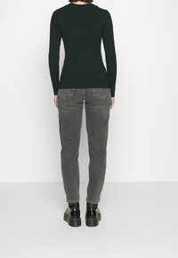 Personne portant un pull noir à côtes, un jean gris décoloré et des bottes noires à lacets, se tenant de dos contre un fond clair.