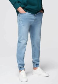 Pantalones jogger de denim azul claro con puños elásticos, que presentan un diseño desgastado y un desgaste discreto. Combinados con zapatillas blancas.