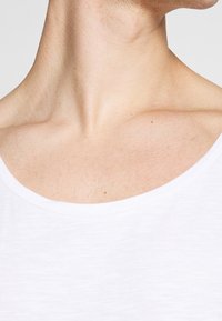 Biała koszulka z szerokim, miękkim dekoltem, odsłaniającym skórę i delikatną teksturę włosów na szyi. Prosta i gładka tkanina.