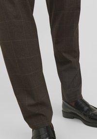 Pantaloni a quadretti marroni realizzati in un tessuto lucido, caratterizzati da un sottile motivo a griglia, abbinati a mocassini neri lucidi.