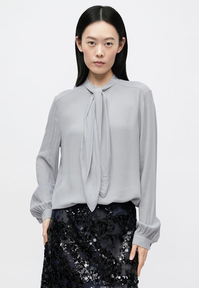 Kobieta o czarnych prostych włosach ubrana w jasnoszarą bluzkę z długim rękawem z wiązaniem z przodu oraz czarną cekinową spódnicę na jednolitym tle.