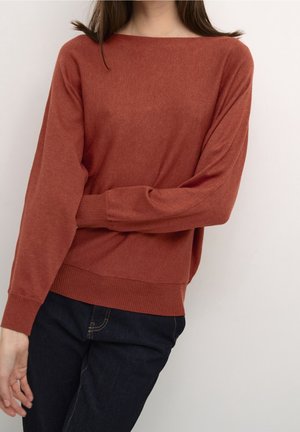 Kvinde iført en rustfarvet langærmet sweater og mørkeblå jeans med armen krydset, stående mod en ensartet lys væg.