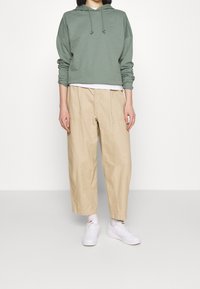 Grön bomullshuva med dragsko, kombinerad med beige vida byxor. Modellen har på sig vita sneakers, vilket visar en avslappnad och ledig outfit.