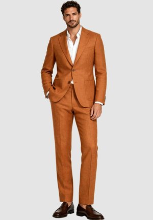 Uomo con capelli scuri e barba che indossa un abito color ruggine, camicia bianca e mocassini marrone scuro, in piedi con una mano in tasca.