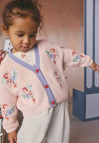 Jauns bērns, kurš valkā rozā džemperi ar ziedu izšuvumiem, violeta apmali, sarkani pogas un svītrainas bikses, stāvot ar nedaudz izstieptām rokām.