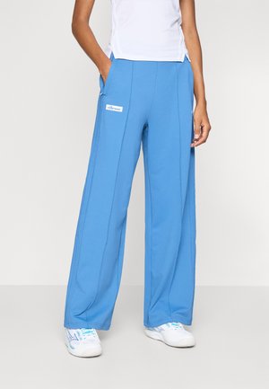 Pantalon large bleu en tissu doux avec des poches latérales et une étiquette de logo blanche sur la cuisse gauche. Associé à des baskets blanches.
