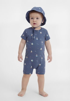 Copil mic purtând un combinezon albastru și o pălărie asortată cu modele mici de bărci cu pânze, stând desculț pe un fundal alb simplu.