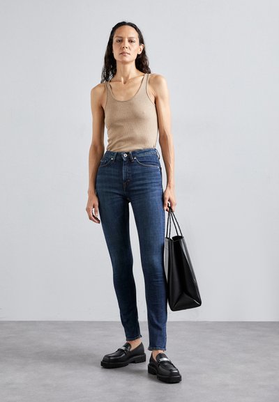 Šviesiai ruda liemenė, aukštu juosmeniu tamsiai mėlyni skinny džinsai, juodi loafersai ir juoda rankinė. Modelis stovi prieš neutralią fono.