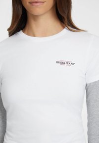 Bílý tričko s krátkým rukávem a kulatým výstřihem, na hrudi s červeným a černým logem "Guess Jeans", pod ním vrstvené šedé dlouhé rukávy.